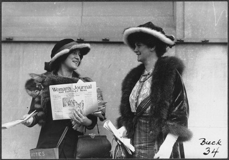 Margaret Foley stands on the right in a fur trimmed hat smiling at the woman on her left who is holding a copy of the Woman's Journal