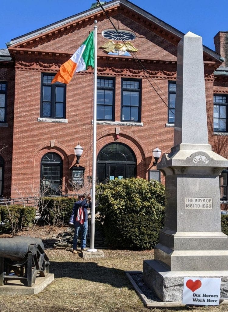The Irish flag is flying in front of the Belfast Maine City Hall, which is a brick building with a gold eagle on the front. 