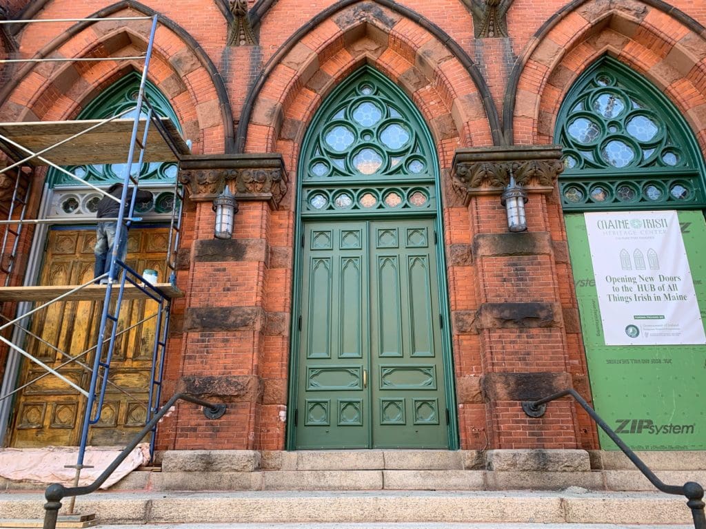 Restoration in progress. Three doors stand at the top of granite steps, left door has scaffolding, middle is freshly painted green, right door holds sign showing sponsors.